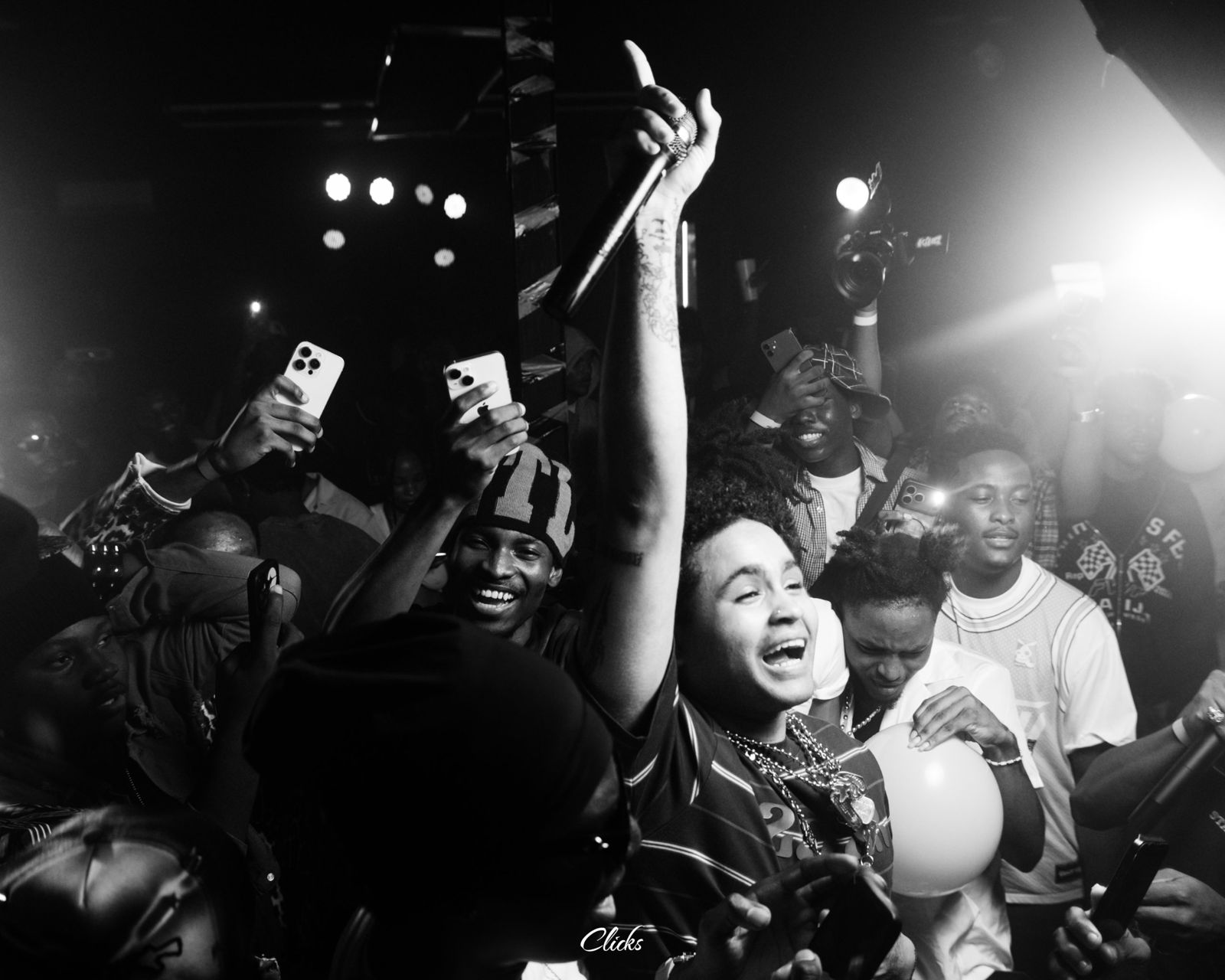 Black and white shot of Suono Sai mid-show, arm raised with a microphone, crowd filming